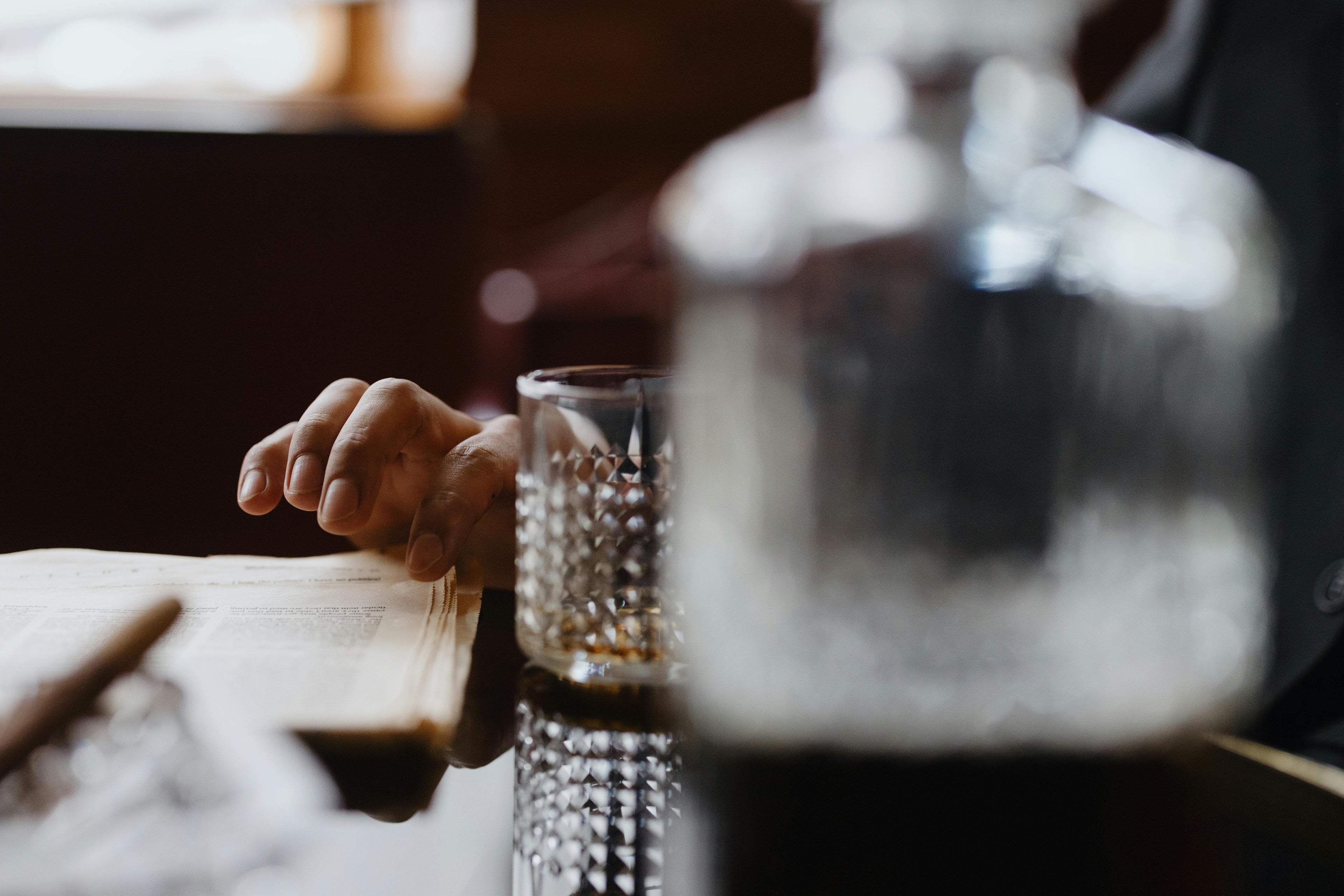 Scatto artistico che immortala un momento di relax in stile vintage: una mano sfoglia un giornale d'epoca mentre un bicchiere di whisky invecchiato e un sigaro acceso completano l'atmosfera sofisticata